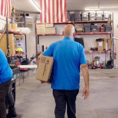 Workers in a garage with an American flag, carrying a box labeled SupplyHouse, next to a branded service vehicle.