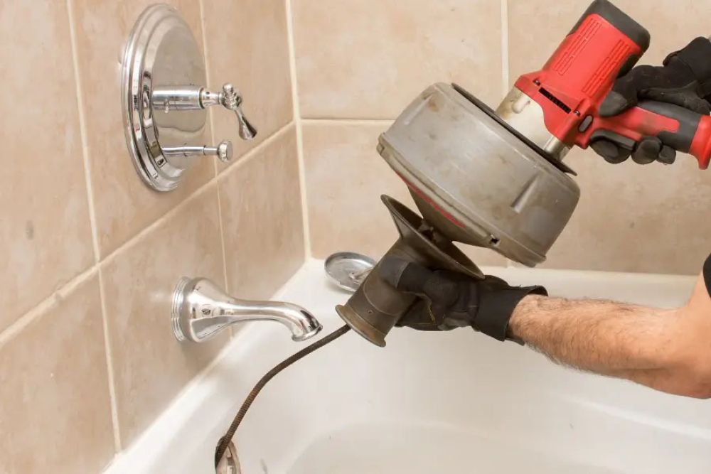Plumber using a drain snake to unclog a bathtub in a tiled bathroom.