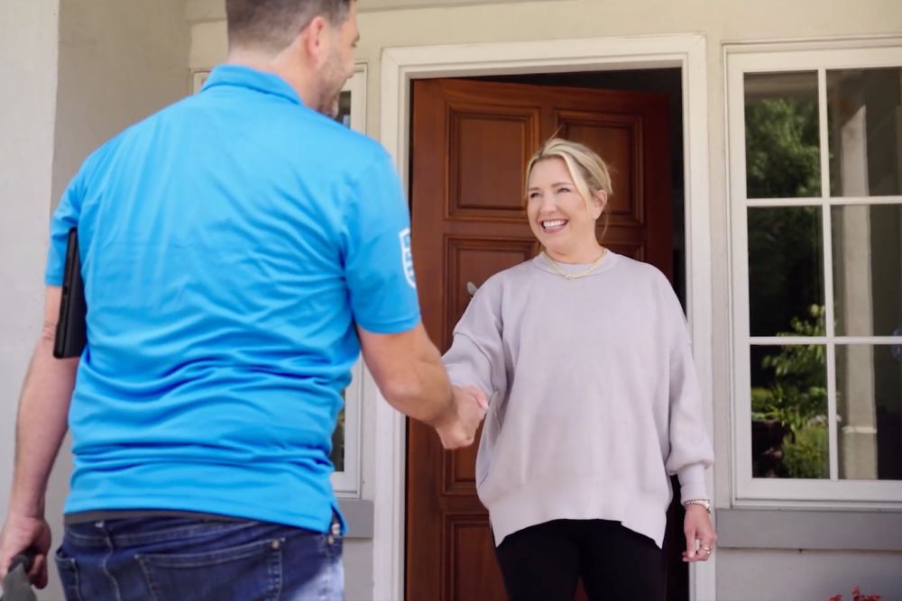 Smiling woman greets repairman at her front door, shaking hands in a friendly exchange.