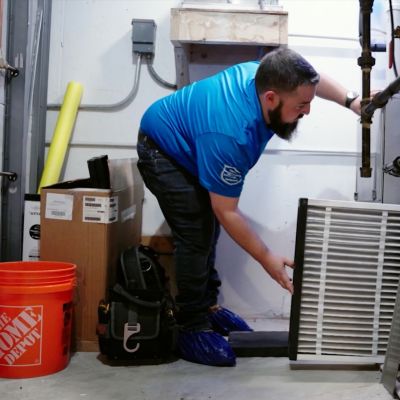 Technician replacing air filter in HVAC system, wearing blue shirt and protective shoe covers.