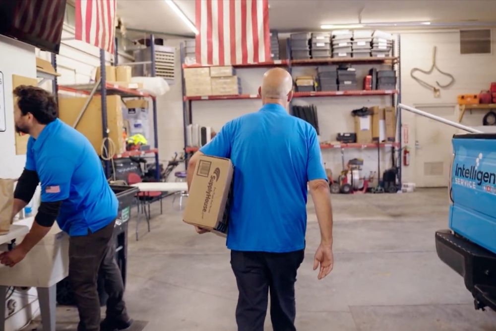 Workers in a garage with an American flag, carrying a box labeled SupplyHouse, next to a branded service vehicle.
