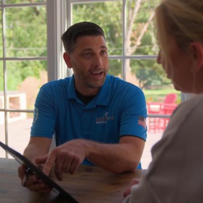 Man in blue uniform explaining tablet to a woman indoors, with bright window view.