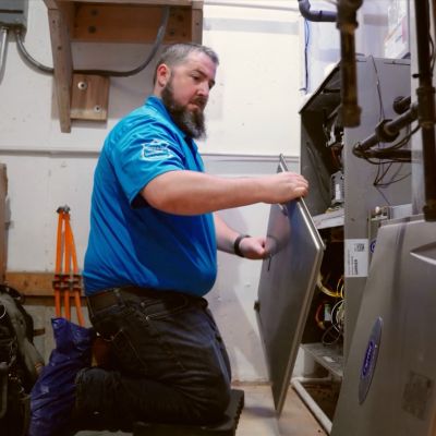 Technician servicing HVAC system in utility room, kneeling and inspecting internal components for maintenance.
