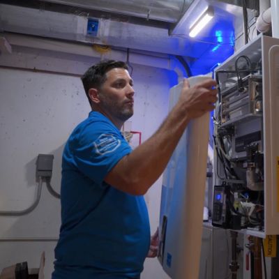 Technician in blue shirt conducting maintenance on a home water heater system in a basement utility room.