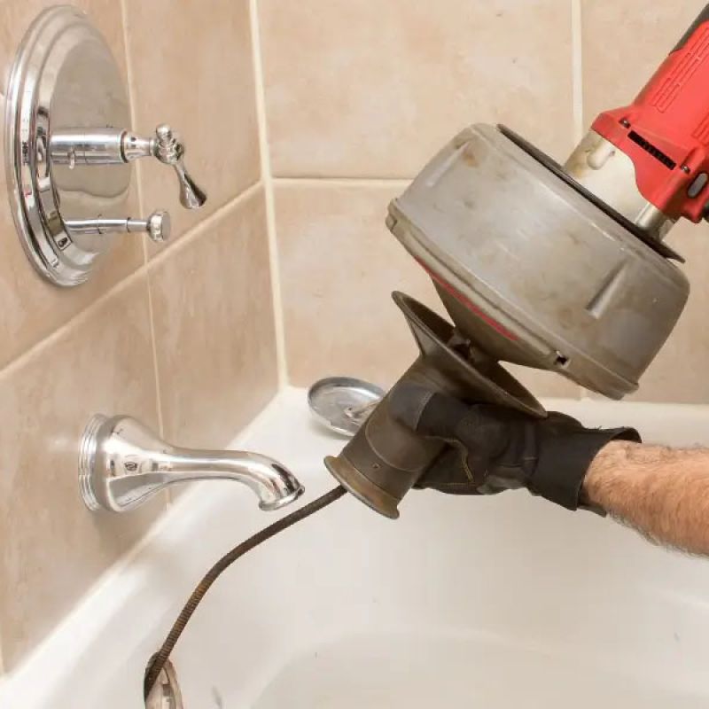 Plumber using a drain snake to unclog a bathtub in a tiled bathroom.