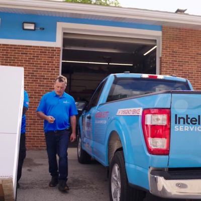 Blue Intelligent Service truck parked outside a garage, with a man in blue shirt walking and a large package nearby.