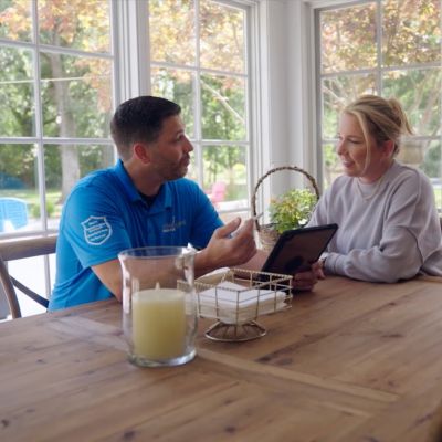 Man in blue shirt consulting a woman with a tablet at a cozy dining table by large windows.
