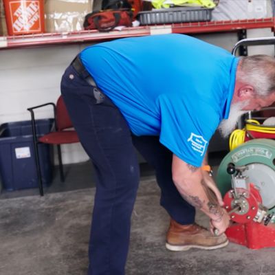 Man using a Spartan tool in a workshop setting, wearing a blue shirt and boots, surrounded by various equipment.